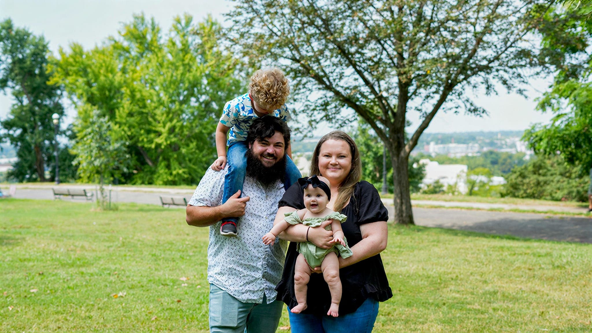 A family of four on a park lawn — a boy on his father's shoulders, a baby in his mother's arms