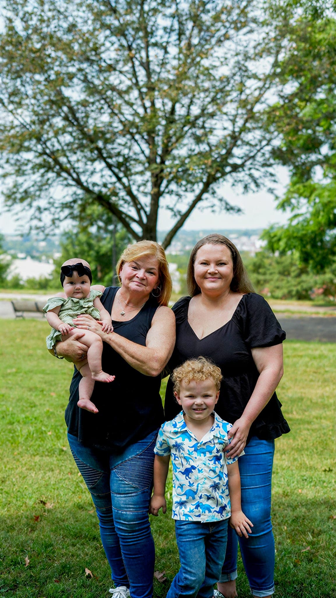 Two women standing on the grass under a tree, one holding a baby, a small boy in front of them
