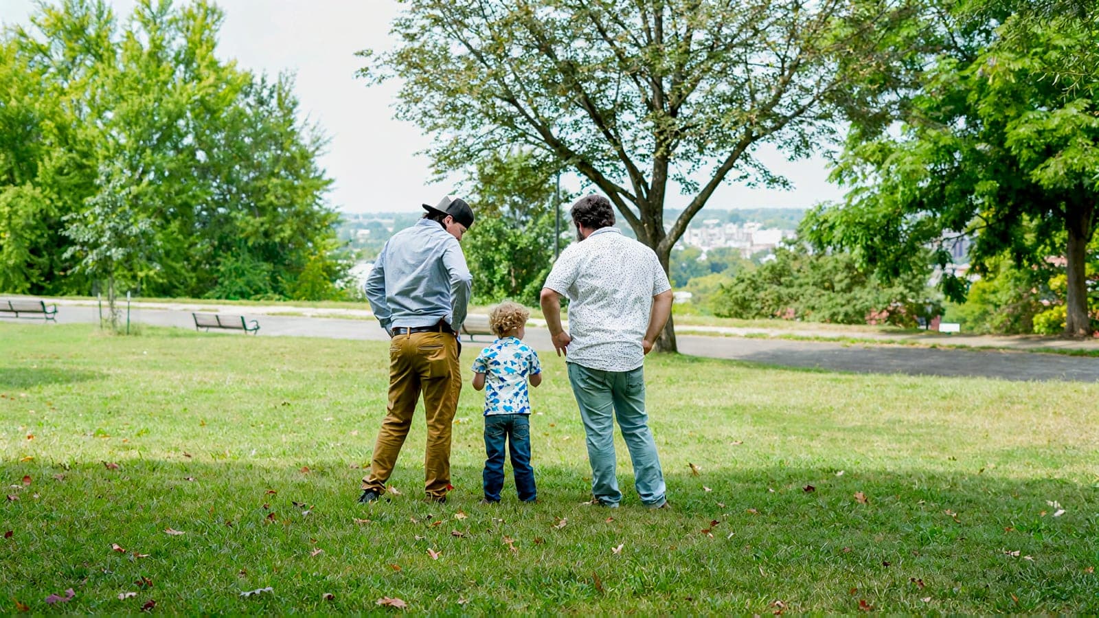 Two men and a small boy standing together on the grass with their backs to the camera, looking out over the city