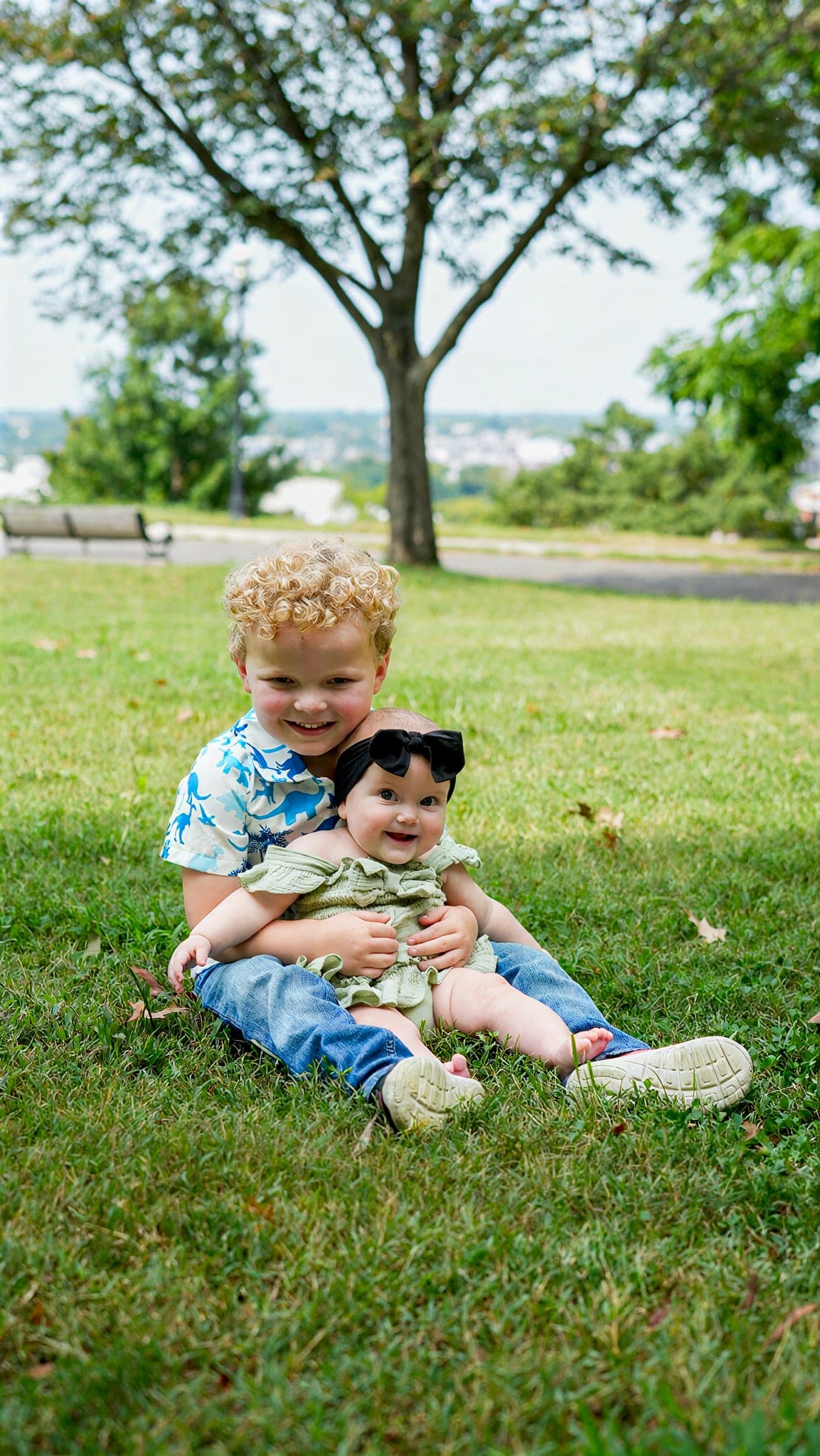 A young boy sitting on a park lawn holding his baby sister, both laughing, with a tree and a view over the city behind them